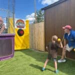 A man sits in a dunk tank as a child throws a ball at the target while an adult guides him. The event is outdoors on artificial grass near a brown shed.
