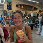 A woman in a green bikini top smiles and holds a shaved ice dessert inside a souvenir shop with shirts, racks, and surfboards in the background.