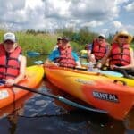 A group of people wearing life jackets paddle rental kayaks on a calm river, surrounded by tall grass and under a partly cloudy sky.