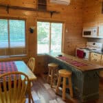 A wooden cabin kitchen with a rustic island, three stools, a dining table with checked tablecloth, stove, microwave, and large windows letting in natural light.