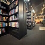 Bookshelves filled with books line a carpeted room with warm lighting. In the background, seating areas and clothing displays are visible.