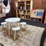 A reading corner with a small round table, four stools, a patterned rug, wooden bookshelves filled with books, and a hanging hammock chair against a wood-paneled wall.