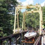 A boat is being transported on a rail track over water using a yellow mechanical lift system surrounded by trees.