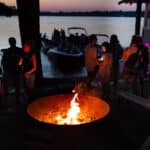 People gather around a fire pit near a lake at dusk, with boats and a house visible in the background.