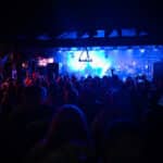 A crowd watches a live band perform on a stage lit with blue and purple lights in a dark indoor venue.