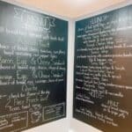 Black chalkboard menu with breakfast and lunch options written in white chalk, including avocado toast, sandwiches, quiche, salads, wraps, kid’s meals, and drinks.