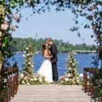 A bride and groom stand together at the end of a flower-lined dock, facing each other by a lake under a clear sky.