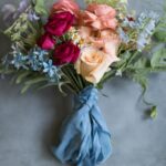 A bouquet of roses and assorted flowers in pink, peach, red, and blue tones, tied with a blue cloth ribbon, displayed on a gray surface.
