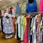 Clothing racks display various colorful dresses, tops, and skirts in a boutique. Wooden floor, white walls, and a mix of casual and formal styles are visible.