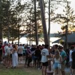 A group of people gather outdoors by a lake at sunset, socializing among tall trees near a cabin or lodge.