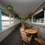 Covered lakeside porch with wooden tables and chairs, potted plants hanging from the ceiling, and large windows overlooking the water.