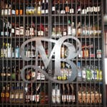 A metal monogram decorates shelves filled with assorted liquor bottles behind a metal grille in a bar, with jackets hanging on the left.