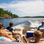 Five people relax on a boat cruising on a lake, surrounded by trees and blue sky, with a cooler visible in the center of the deck.
