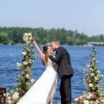 A bride and groom kiss outdoors on a dock by a lake, surrounded by floral arrangements, under a clear blue sky.