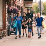 A group of six people, including kids and adults, walk together on a sidewalk in a small town, carrying shopping bags.