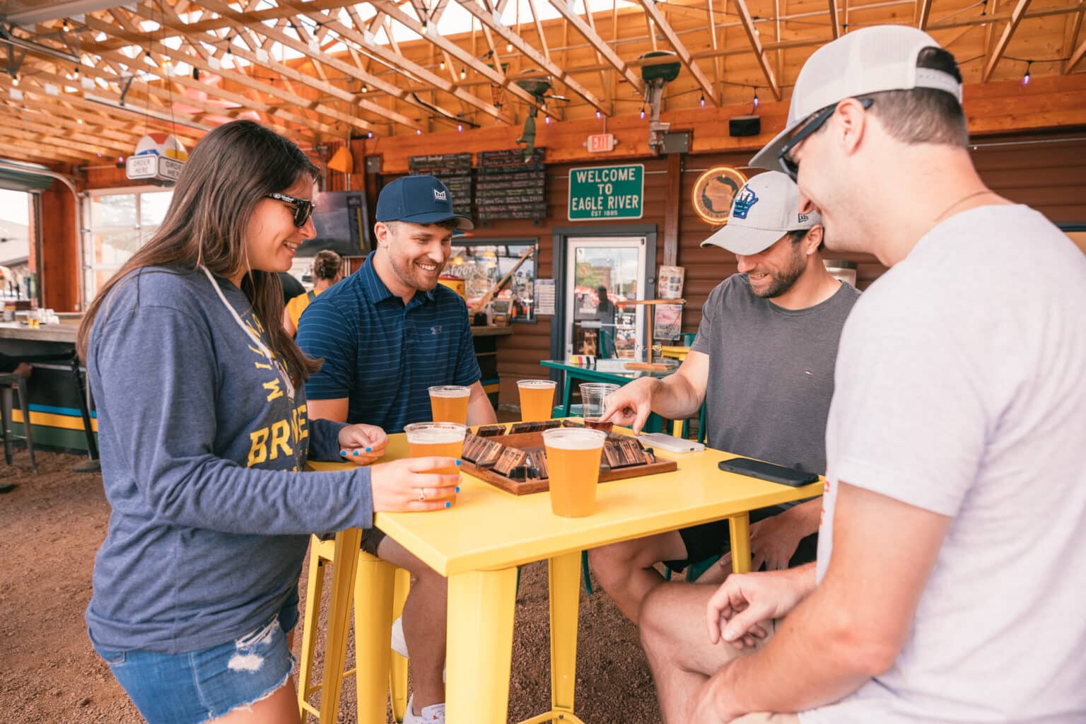 Four people sit around a yellow table with drinks, playing a board game inside a wooden, open-air bar or restaurant.