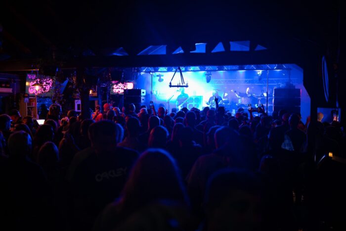 A crowd watches a live band perform on a brightly lit stage in a dark indoor venue.