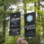 Two black signs on a pole advertise Marina Bar event parking and The Everett Resort, with a hanging basket of pink flowers below, surrounded by green trees.