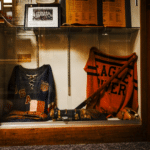 Display case with vintage sports jerseys, bats, gloves, and photos, including one jersey labeled “USA” and another labeled “Eagle River.”.