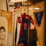 A display case contains a 1988 USA Hockey Team pennant, a blue and red hockey jersey with number 55, a blue Suomi Finland pennant, a portrait sketch, and several photos.