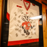 Framed, autographed University of Wisconsin Badgers hockey jersey displayed on a wooden wall.