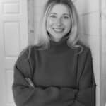 Woman with long blonde hair wearing a turtleneck sweater stands indoors, smiling with arms crossed in front of a paneled wall.