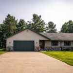Single-story house with a two-car garage, stone and siding exterior, concrete driveway, and a well-kept lawn bordered by trees.