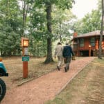 Two people walk on a path toward a brown cabin in a wooded area, with a blue vehicle parked nearby and a mailbox labeled 3802 visible.