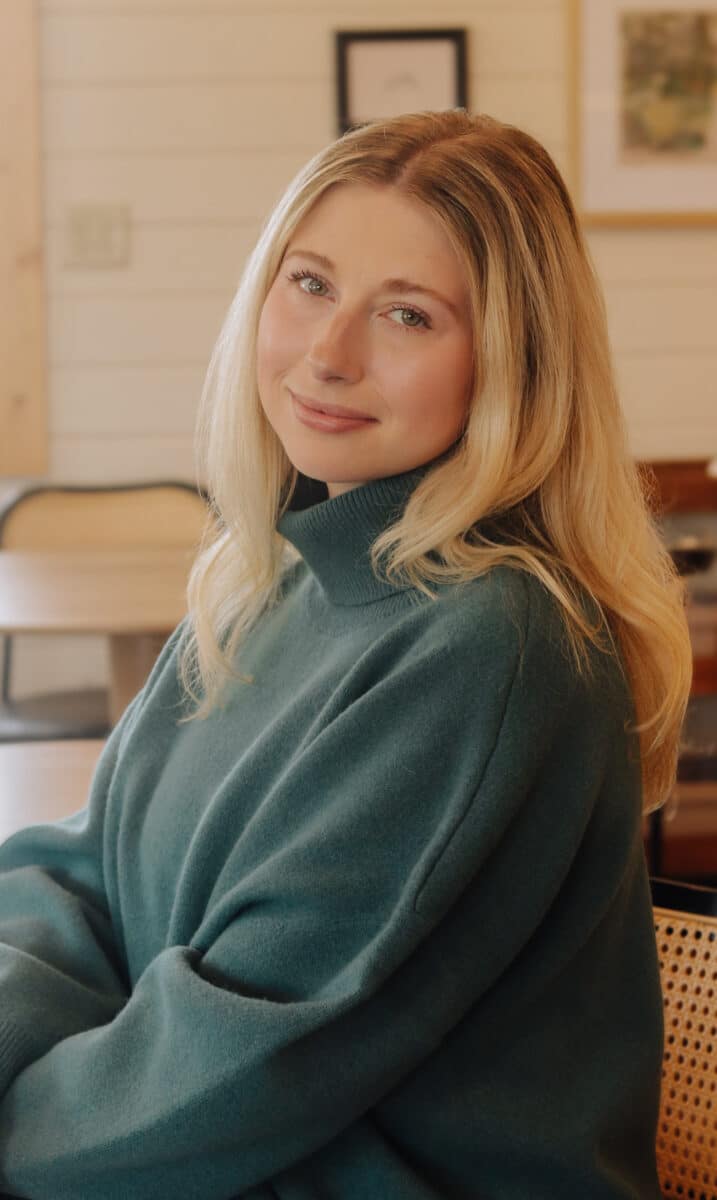 Hannah Hallstrom Polich Shorewest Realtors A woman with long blonde hair wearing a teal turtleneck sweater sits indoors, looking at the camera with a slight smile.