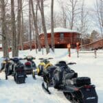 Several snowmobiles are parked in the snow near a group of red wooden cabins surrounded by leafless trees on a winter day.