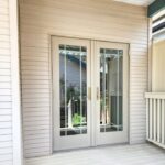 Double glass doors with gold handles on a beige deck, surrounded by beige siding and partially enclosed by a railing. Reflections of plants and a house are visible in the glass.