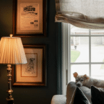 A small dog rests on a couch by a window; nearby are a lit table lamp and framed wall art, including a Dalmatian drawing.