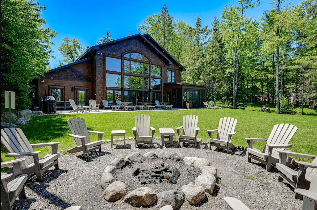 A brown house with large windows overlooks a lawn with a stone fire pit surrounded by Adirondack chairs.