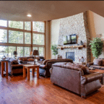 Living room with multiple leather sofas, a stone fireplace, and a large wall of windows overlooking a lake.