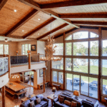 A multi-level living area with exposed wooden beams, a large antler chandelier, and floor-to-ceiling windows overlooking a lake.