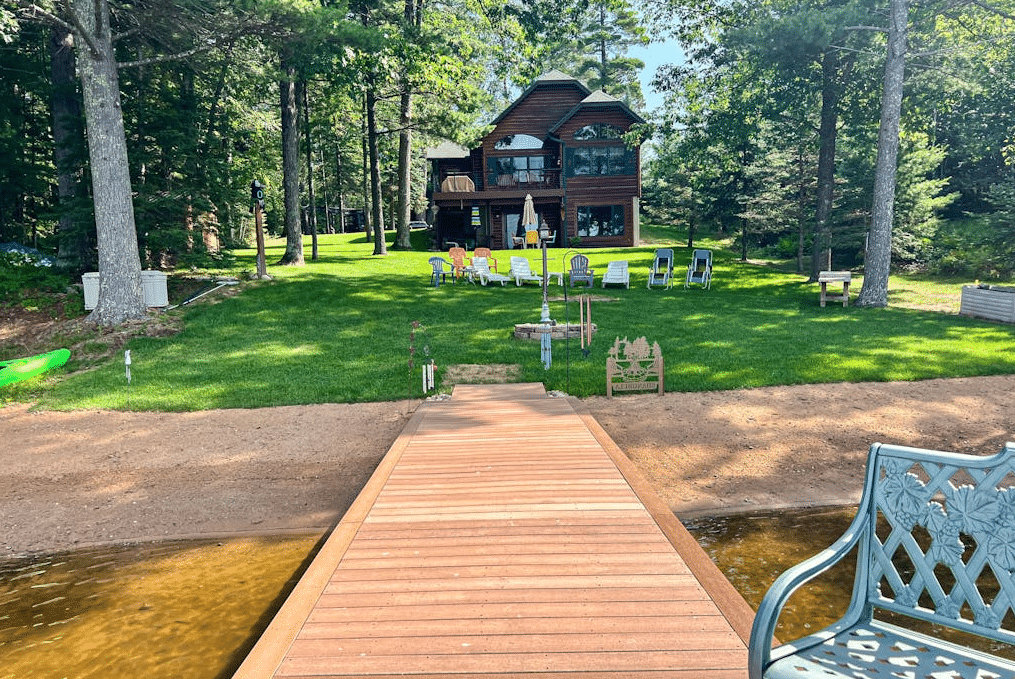 A wooden dock leading to a lawn with several chairs and a multi-story cabin in the background.
