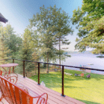 A wooden deck with metal furniture overlooking a grassy lawn, trees, and a lake.