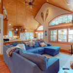 Open-plan living room with a large blue sectional sofa, a stone fireplace, and a kitchen area with wood ceilings and flooring.