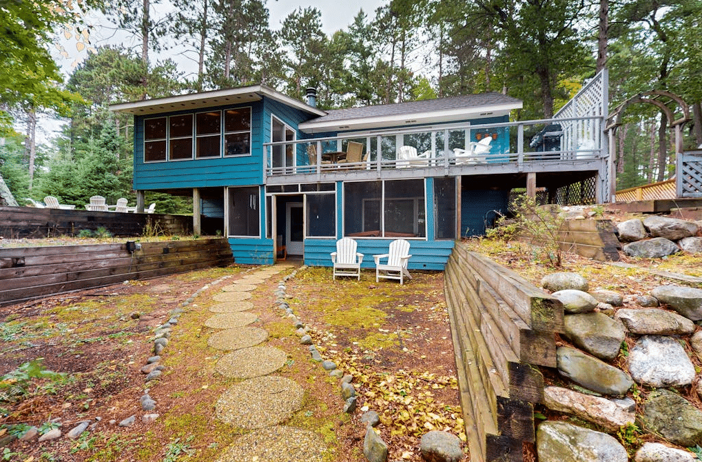 4a A two-story blue house in a forest with a second-floor deck, a screened-in porch on the lower level, and a stone path leading to two white chairs in the yard.