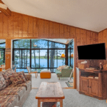 Living room with wood paneled walls, a patterned sofa, wooden coffee table, and flat-screen TV, looking out through large windows at a lake.
