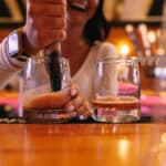 A bartender uses a muddler to mix a drink in a glass on a bar next to a glass of whiskey.