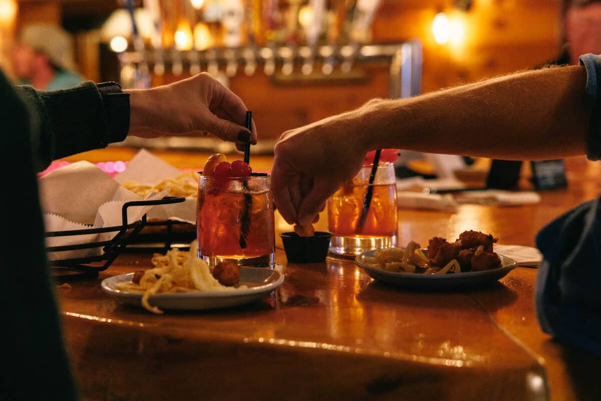 ER_Kickback_20260930_4591 Two people at a bar table with cocktails and plates of fried food.
