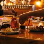 Two people at a bar table with cocktails and plates of fried food.