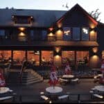A two-story building at dusk with a large outdoor deck featuring tables, chairs, and red umbrellas.