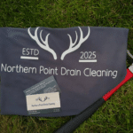 A grey Northern Point Drain Cleaning banner with a logo of deer antlers, featuring two business cards and a tool with a red handle lying on grass.