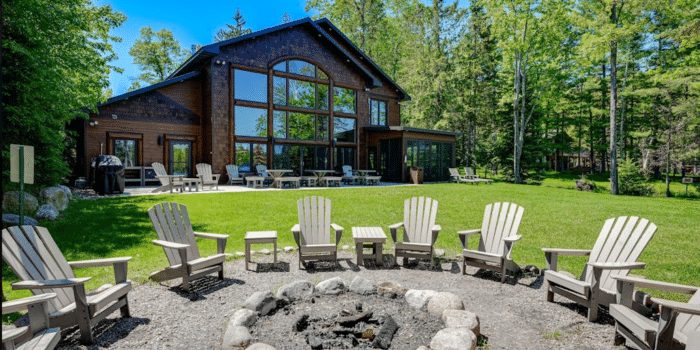 A brown house with large windows overlooks a lawn with a stone fire pit surrounded by Adirondack chairs.