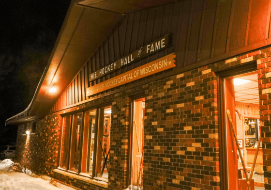 Brick building labeled “WIS. Hockey Hall of Fame” at night, with snow on the ground and light coming from large windows displaying hockey memorabilia.