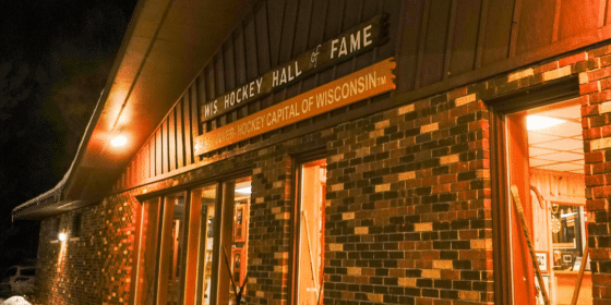 Brick building labeled “WIS. Hockey Hall of Fame” at night, with snow on the ground and light coming from large windows displaying hockey memorabilia.