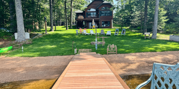 A wooden dock leading to a lawn with several chairs and a multi-story cabin in the background.