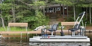 A small pontoon boat with two seats floats near a wooden dock and benches, with a cabin and steps surrounded by trees in the background.